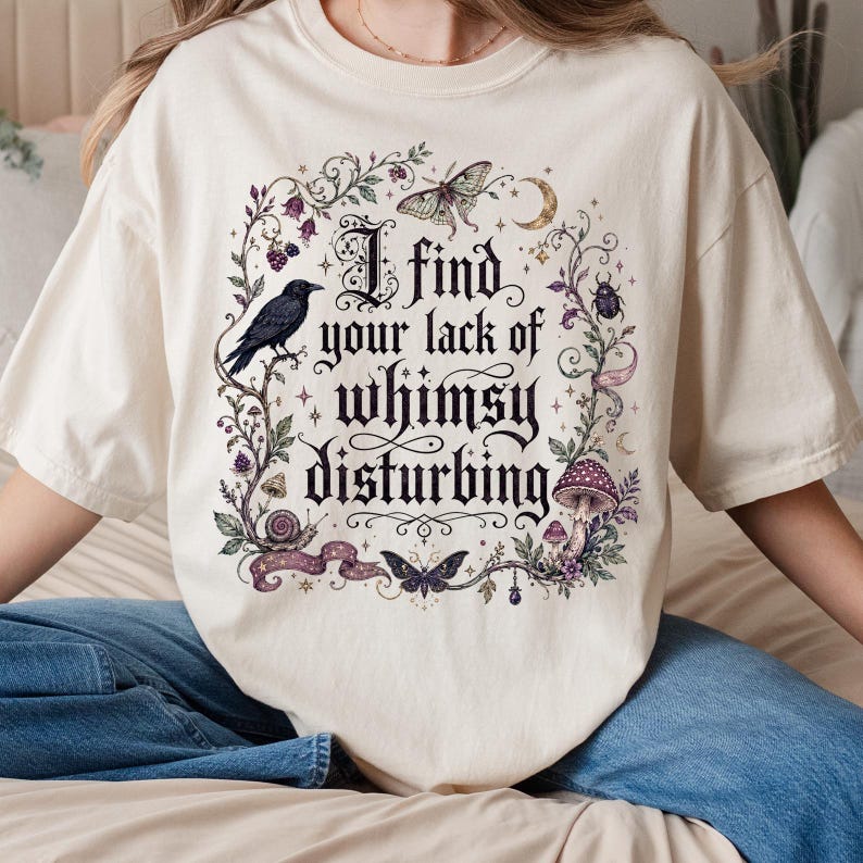 May include: A cream-colored short-sleeve t-shirt featuring a central graphic with the text "I find your lack of whimsy disturbing." The design includes a black raven, a moth, a snail, a beetle, a mushroom, and floral vines in dark tones.