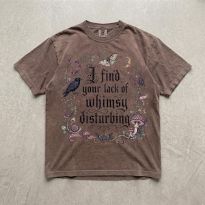 May include: A brown short-sleeve t-shirt featuring a graphic print with the text "I find your lack of whimsy disturbing." The design includes illustrations of a black crow, a moth, a butterfly, a mushroom, and floral vines in purple and green.
