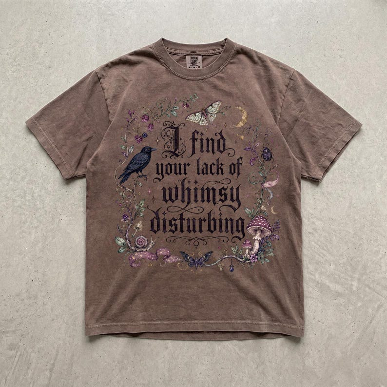 May include: A brown short-sleeve t-shirt featuring a graphic print with the text "I find your lack of whimsy disturbing." The design includes illustrations of a black crow, a moth, a butterfly, a mushroom, and floral vines in purple and green.