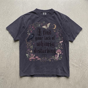 May include: A charcoal gray short-sleeve t-shirt featuring a central graphic with the text 'I find your lack of whimsy disturbing' surrounded by illustrations of a crow, a moth, a crescent moon, mushrooms, vines, and small floral elements.