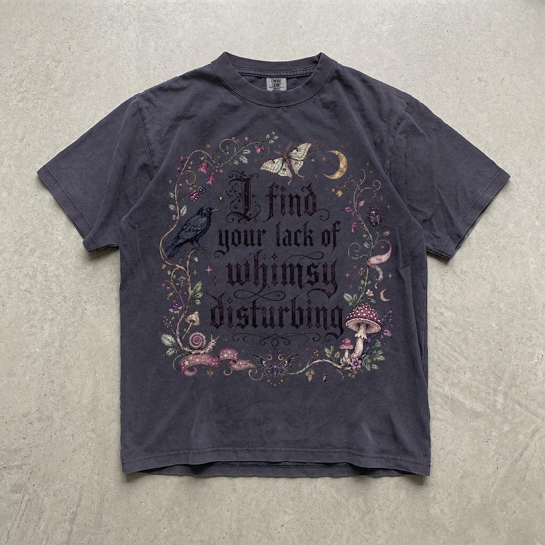 May include: A charcoal gray short-sleeve t-shirt featuring a central graphic with the text 'I find your lack of whimsy disturbing' surrounded by illustrations of a crow, a moth, a crescent moon, mushrooms, vines, and small floral elements.