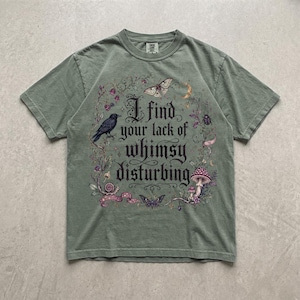 May include: A sage green short-sleeve t-shirt featuring a graphic print with the text "I find your lack of whimsy disturbing." The design includes a black crow, a moth, a snail, a mushroom, a beetle, and floral elements in shades of purple and brown.