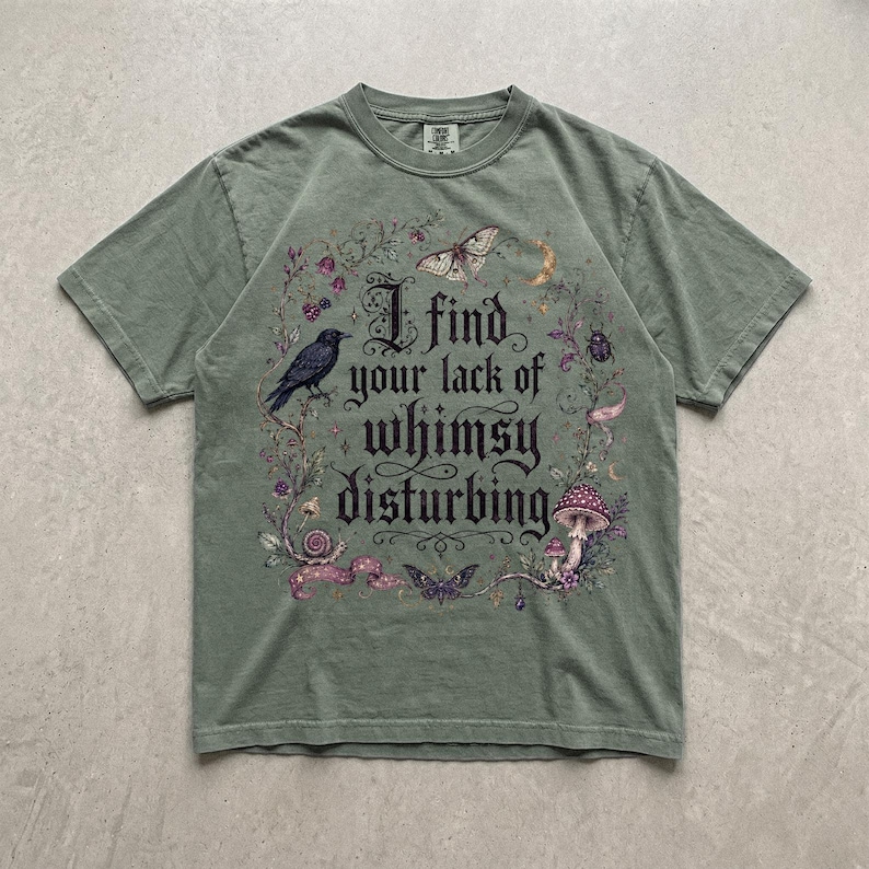 May include: A sage green short-sleeve t-shirt featuring a graphic print with the text "I find your lack of whimsy disturbing." The design includes a black crow, a moth, a snail, a mushroom, a beetle, and floral elements in shades of purple and brown.