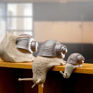May include: Three decorative snail ornaments. The snails have beige bodies and metallic silver shells. One snail sits upright, while the other two appear to be crawling over a wooden ledge. The background is blurred.