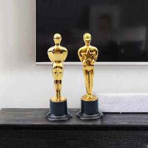 May include: Two gold-coloured statuettes, one holding a star, stand on a dark surface. The figures have a classic design with a smooth, reflective finish. Each is mounted on a black base, creating a striking contrast.