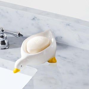 May include: A white ceramic soap dish in the shape of a goose, featuring yellow feet and beak. It holds a bar of soap. The soap dish is on a marble countertop, with a chrome tap in the background.