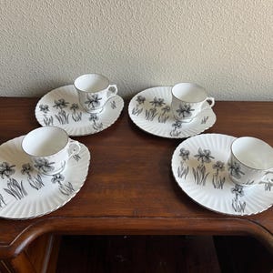 May include: A set of four white porcelain tea cups and matching scalloped plates, each decorated with a black and white botanical iris flower illustration. The cups feature a curved handle and the plates have a ribbed texture along the edges.