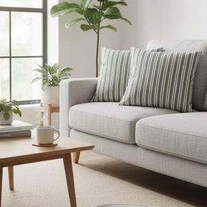 Puede incluir: Un sofá gris claro con dos cojines a rayas y un cojín de suelo a juego. Los cojines tienen un patrón de rayas verdes y blancas. También se ven una mesa de centro de madera y una planta en maceta.