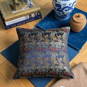May include: A patterned cushion with a dark blue base and gold and red detailing. It sits on a blue mat, alongside a blue and white ceramic jar, a small woven basket, and books. A gold lantern is also visible in the background.