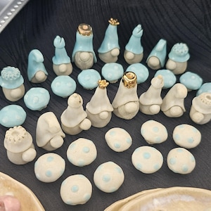 May include: A collection of ceramic gnomes and mushroom-shaped figurines. The gnomes have light blue hats and some have gold accents. The mushrooms are white with light blue polka dots. The figurines are arranged on a dark surface.