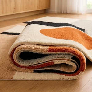 May include: A plush area rug featuring a geometric pattern with abstract shapes in burnt orange, cream, black, and tan. The rug is partially rolled, revealing a thick, textured pile and a layered design that combines solid color blocks with curved forms.