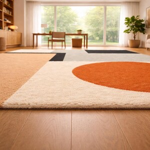 May include: A rectangular area rug featuring a geometric pattern with a large orange circle, a light beige section, a white field, and black accents. The rug has a textured, tufted pile surface and is laid flat on a light brown wooden floor.