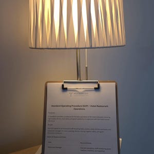 May include: A white pleated lampshade sits atop a silver lamp base, illuminating a clipboard with a document titled "Standard Operating Procedure (SOP) - Hotel Restaurant Operations". The document includes text about restaurant procedures.