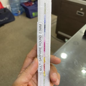 May include: A white rectangular display card with a vertical row of multi-colored round sapphires. The sapphires are arranged in a gradient of colors, from green to yellow to pink to blue. The card has the text "MULTI SAPPHIRE ROUND 2.5MM".