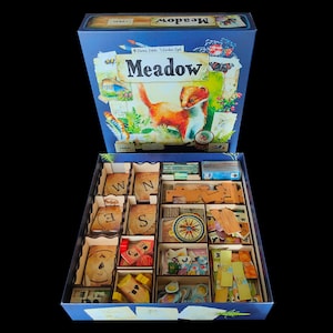 Op de afbeelding: Een bordspeldoos met de titel "Meadow", geopend om de spelcomponenten te onthullen. De dooskunst toont een vos en natuurscènes. Binnenin zijn spelonderdelen gerangschikt in compartimenten, waaronder kaarten en houten fiches.