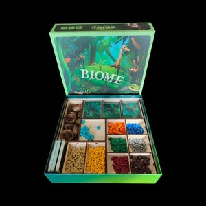 Könnte beinhalten: Geöffnete Brettspielbox mit dem Titel "BIOME" und einer Dschungelszene auf dem Deckel. Im Inneren sind verschiedene Spielsteine in Fächern angeordnet, darunter Holzmarker, farbige Würfel und Karten. Die Box ist grün.