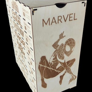 Peut inclure: Boîte en bois avec des illustrations Marvel gravées au laser. La boîte présente un design de Spider-Man accroupi et le logo Marvel. Les côtés affichent des noms de personnages comme Black Panther et Thor. La boîte est en bois clair.