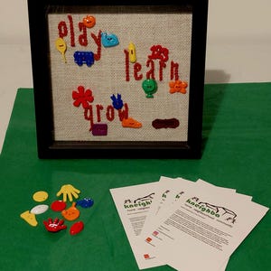 以下が含まれることがあります： 「play, learn, grow」の文字が赤い糸で刺繍された、ジュートの背景に額装されたアートワーク。カラフルで遊び心のある形やオブジェクトがジュートに取り付けられています。いくつかの印刷された紙が緑色の表面にあります。