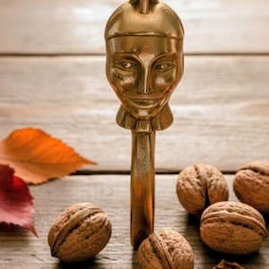 Könnte beinhalten: Ein Messing-Nussknacker in Form eines Gesichts steht inmitten mehrerer Walnüsse, von denen einige geöffnet sind. Der Nussknacker steht auf einer Holzoberfläche mit Herbstblättern.