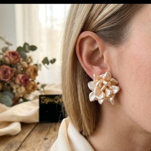 Könnte beinhalten: Nahaufnahme eines Ohres einer Frau, das mit einem großen, hellfarbenen floralen Ohrstecker geschmückt ist. Der Ohrring hat mehrere blütenblattartige Schichten. Im Hintergrund ist ein Strauß getrockneter Blumen zu sehen, zusammen mit einer schwarzen Karte.
