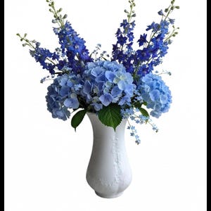 Könnte beinhalten: Eine weiße Keramikvase mit einem dezenten Reliefmuster enthält ein großes Arrangement aus blauen Hortensienblüten, hohen Stielen dunkelblauer Rittersporne und kleinen Büscheln hellblauer Vergissmeinnicht mit grünen Blättern.