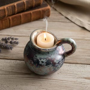 Könnte beinhalten: Ein kleiner, kugelförmiger Kerzenhalter aus Keramik mit Griff, glasiert in Blau-, Grün- und Lilatönen. Eine brennende weiße Kerze befindet sich im Inneren, mit aufsteigendem Rauch. Der Halter steht auf einer Holzoberfläche, mit Büchern und Stoff im Hintergrund.