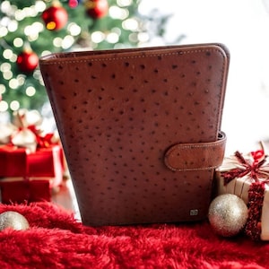 Könnte beinhalten: Ein brauner Lederplaner mit strukturierter Oberfläche und Druckknopfverschluss. Der Planer befindet sich auf einem roten Teppich, mit Weihnachtsdekorationen und verpackten Geschenken im Hintergrund.
