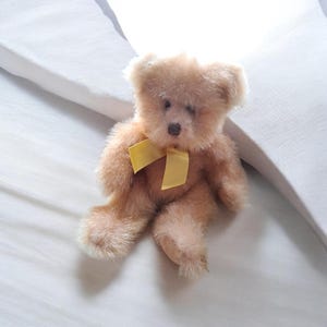 Könnte beinhalten: Ein kleiner, hellbrauner Teddybär mit einer gelben Schleife um den Hals. Das Plüschtier hat eine dunkelbraune Nase und sitzt auf einer weißen Oberfläche. Das Fell des Bären ist weich und flauschig.