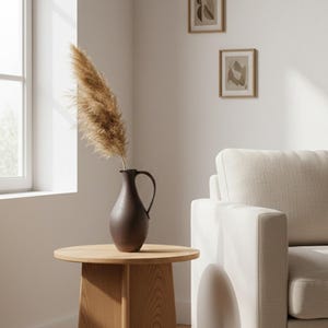Könnte beinhalten: Eine dunkelbraune Vase mit Henkel enthält getrocknetes Pampasgras, das auf einem runden Beistelltisch aus Holz steht. Zwei gerahmte abstrakte Kunstwerke hängen an der weißen Wand. Im Hintergrund befindet sich ein cremefarbenes Sofa.