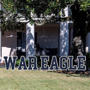 May include: Large, navy blue and white letters spell "WAR EAGLE" on a green lawn in front of a white building with columns and a tree. The building has a balcony and a front door.