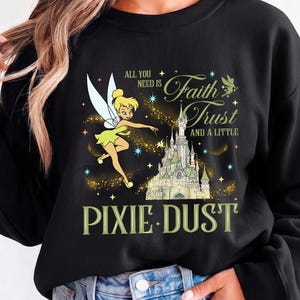 Pode incluir: Uma camisola preta de manga comprida com um gráfico da Sininho a voar sobre um castelo. O texto diz: All you need is Faith Trust and a little Pixie Dust. O design inclui detalhes em amarelo, branco e azul com estrelas cintilantes.
