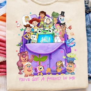 May include: A cream-colored t-shirt featuring a graphic of a purple backpack filled with various cartoon characters from the Toy Story franchise. The text "YOU'VE GOT A FRIEND IN ME" is printed in pink below the backpack illustration.