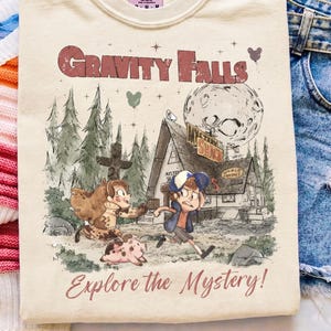 May include: A cream-colored t-shirt featuring a graphic print of the Gravity Falls Mystery Shack, two cartoon characters, and a pig. The text reads Gravity Falls and Explore the Mystery. The shirt has a Comfort Colors brand label at the neck.