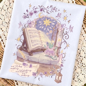 May include: A light blue t-shirt featuring a graphic illustration of a green lizard in a dress standing before an open book on a stand. The scene includes a window, stars, candles, a quill, and the text, "and I'll keep wondering and wondering and wondering and wondering..."