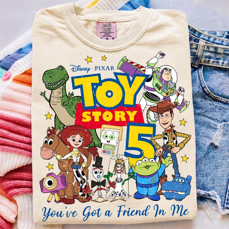 May include: A cream-colored t-shirt featuring a graphic print of Toy Story characters including Woody, Buzz Lightyear, Jessie, Rex, and others. The design includes the text Toy Story 5 and the phrase You've Got a Friend in Me in blue lettering.