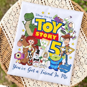 May include: A white t-shirt featuring a colorful Toy Story 5 graphic with characters including Woody, Buzz Lightyear, Jessie, Bullseye, Rex, and others. The design includes the text Toy Story 5 and the phrase You've Got a Friend In Me.