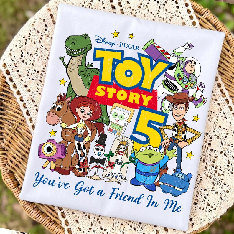 May include: A white t-shirt featuring a colorful Toy Story 5 graphic with characters including Woody, Buzz Lightyear, Jessie, Bullseye, Rex, and others. The design includes the text Toy Story 5 and the phrase You've Got a Friend In Me.