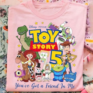 May include: A light pink t-shirt featuring a colorful graphic of Disney Pixar Toy Story characters including Rex, Buzz Lightyear, Woody, Jessie, Bullseye, and others. The text reads Toy Story 5 and You've Got a Friend In Me.