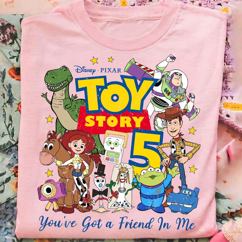 May include: A light pink t-shirt featuring a colorful graphic of Disney Pixar Toy Story characters including Rex, Buzz Lightyear, Woody, Jessie, Bullseye, and others. The text reads Toy Story 5 and You've Got a Friend In Me.