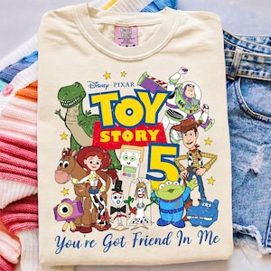 Könnte beinhalten: Ein cremefarbenes T-Shirt mit einer bunten Grafik der Toy Story-Figuren, darunter Woody, Buzz Lightyear, Rex, Jessie und Bullseye. Das Design enthält den Text Toy Story 5 sowie You've Got Friend In Me in Blau und Gelb.