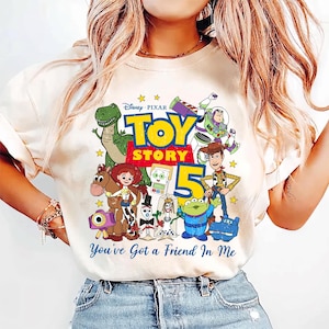 May include: A cream-colored t-shirt featuring a colorful graphic of Toy Story characters including Woody, Buzz Lightyear, Jessie, Rex, and others. The design includes the text Disney Pixar Toy Story 5 and You've Got a Friend In Me.