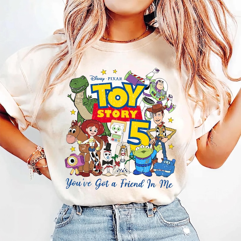 May include: A cream-colored t-shirt featuring a colorful graphic of Toy Story characters including Woody, Buzz Lightyear, Jessie, Rex, and others. The design includes the text Disney Pixar Toy Story 5 and You've Got a Friend In Me.