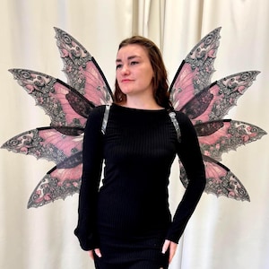 May include: A person wearing a black long-sleeve dress with a thigh-high slit, paired with patterned tights. The person is wearing large, decorative fairy wings with pink and black details.