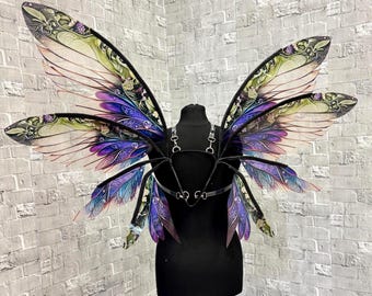 May include: A pair of fantasy-themed fairy wings with a black harness. The wings feature a gradient of colors, including purple, green, and pink, with intricate floral patterns. The wings are displayed on a black dress form against a brick wall.
