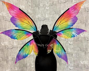 May include: Rainbow-colored fairy wings with a black harness, displayed on a black mannequin. The wings feature vibrant hues of red, orange, yellow, green, blue, and purple. The wings are attached to a black harness with silver-colored hardware.