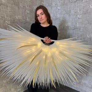 May include: A large, illuminated white and gold decorative wing structure. The wings are held by a person wearing a black top and pants. The wings have a feathery appearance with gold glitter accents and small lights.