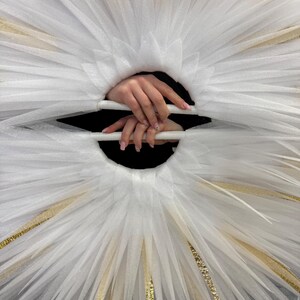 May include: A white tulle halo-like structure with gold accents. The design features radiating layers of fabric, creating a starburst effect. The center reveals a dark void, with hands holding the structure.