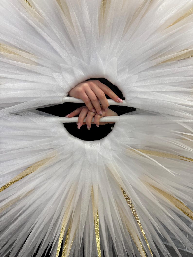 May include: A white tulle halo-like structure with gold accents. The design features radiating layers of fabric, creating a starburst effect. The center reveals a dark void, with hands holding the structure.