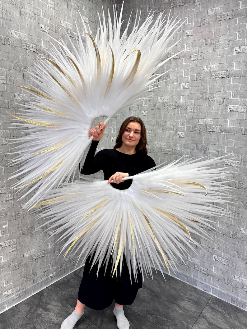 May include: Two large, white feather fans with gold accents. The fans are held open, creating a symmetrical, circular shape. The feathers are long and pointed, radiating outwards from a central handle. The gold accents are curved and add a touch of elegance.