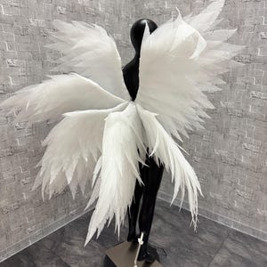 Może przedstawiać: Duże białe skrzydła anioła z warstwowym wzorem piór, wystawione na czarnym manekinie. Skrzydła są szeroko rozłożone, tworząc dramatyczny efekt. Manekin stoi na czarnej podstawie ze srebrnym stojakiem.
